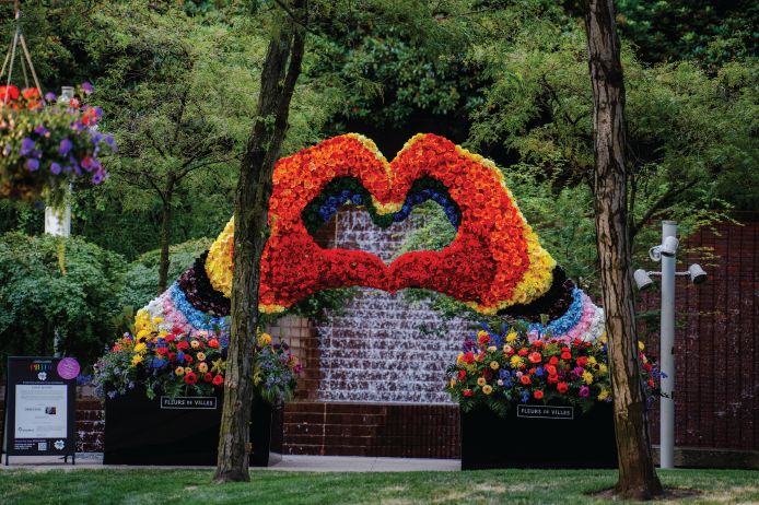 Large, oudoor sculpture of hands making the shape of a heart. The hands are covered in flowers in the colours of the Pride flag.