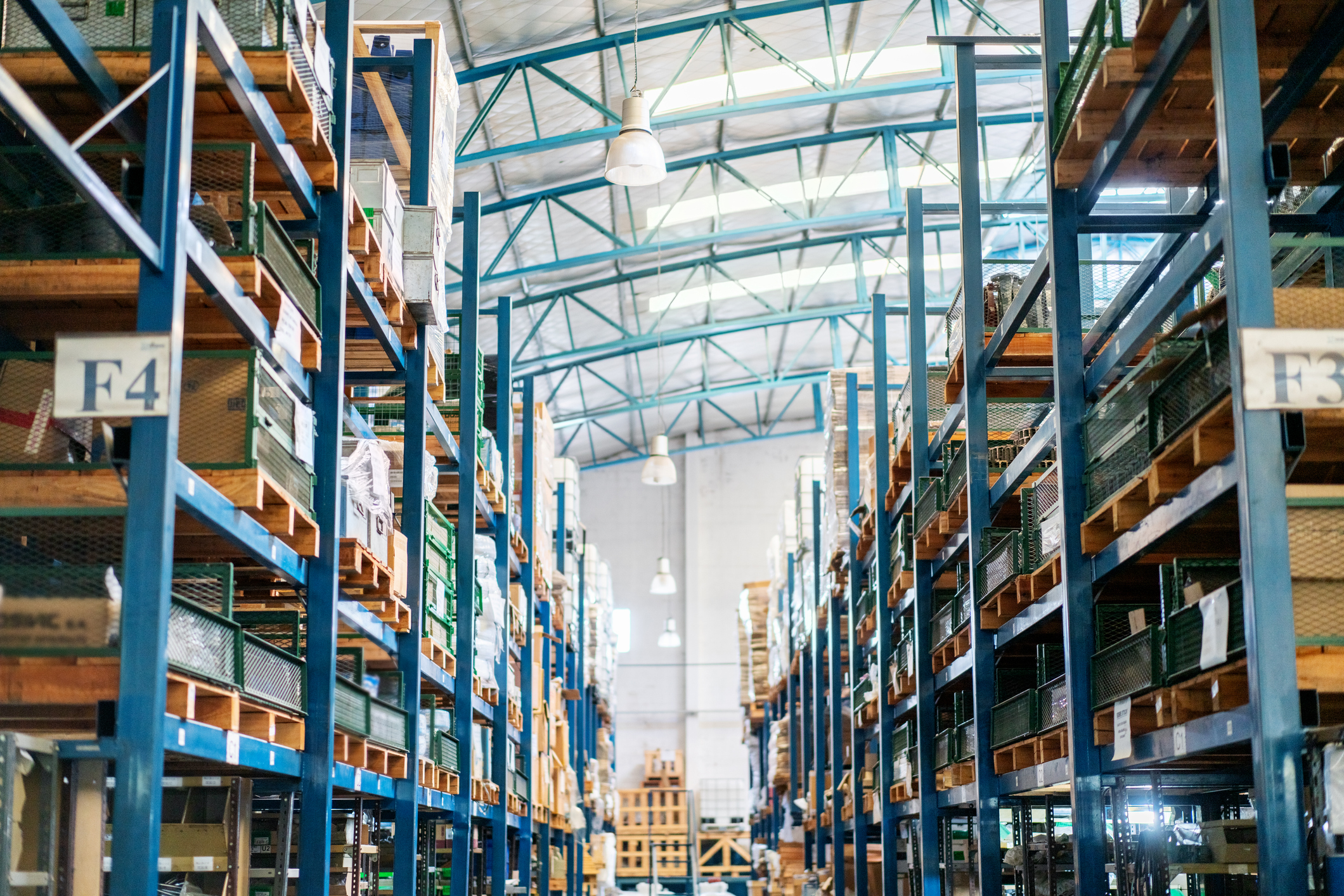 Interior of a large warehouse with tall blue metal shelves filled with various boxes and items. The shelves are organized in rows, and there are signs indicating sections such as "F4" and "F3". The warehouse has a high ceiling with metal beams and several hanging lights.