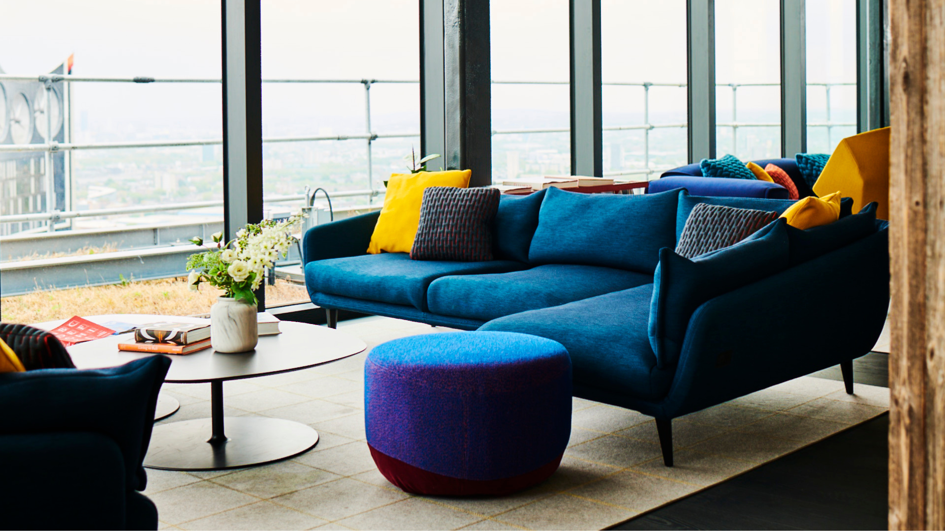 Modern living room with a blue sectional sofa, colorful pillows, coffee table, and city view through large windows.