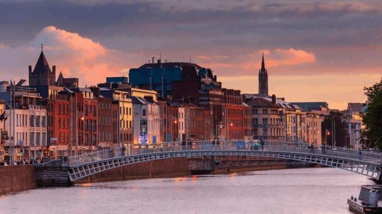 A scenic city view at sunset, featuring a pedestrian bridge over a river, colorful buildings along the waterfront, and historic spires in the background.