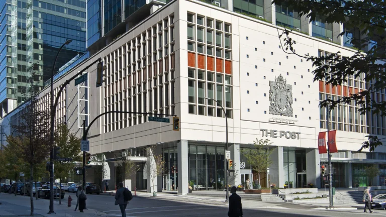 Street view of The Post building in downtown Vancouver, featuring a modern facade with vertical window panels, the building name ‘The Post’ and a large crest on the exterior wall. Pedestrians are crossing the intersection in front, and surrounding high-rise glass towers are visible in the background.