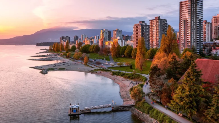Vue du front de mer de Vancouver au coucher du soleil : plage de sable, promenade arborée, immeubles et montagnes en arrière-plan.