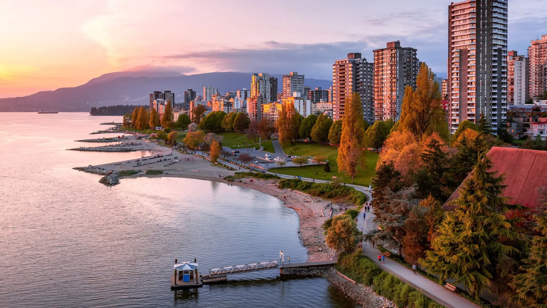 Vue du front de mer de Vancouver au coucher du soleil : plage de sable, promenade arborée, immeubles et montagnes en arrière-plan.