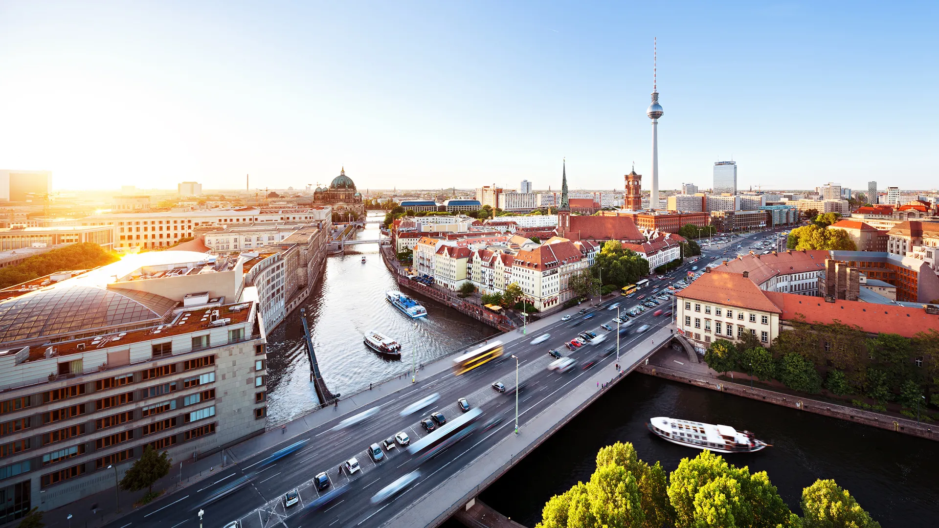Panoramic view of Berlin cityscape featuring the Spree River, bridges, boats, historic buildings, and the Berlin TV Tower under a clear blue sky.