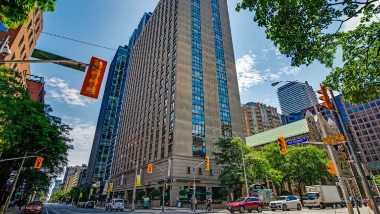 Street‑level view of a tall modern high‑rise surrounded by other buildings, trees, traffic lights, and cars at a downtown intersection.