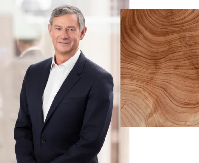 A professionally dressed person in a dark blazer and light shirt stands against a softly blurred indoor background. The person is positioned on the left side of the image, facing forward. On the right side, a rectangular overlay shows a close-up of wood grain with visible rings and warm brown tones.