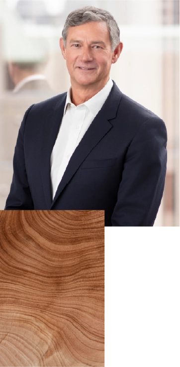 A professionally dressed person in a dark blazer and light shirt stands against a softly blurred indoor background. The person is positioned on the left side of the image, facing forward. On the right side, a rectangular overlay shows a close-up of wood grain with visible rings and warm brown tones.