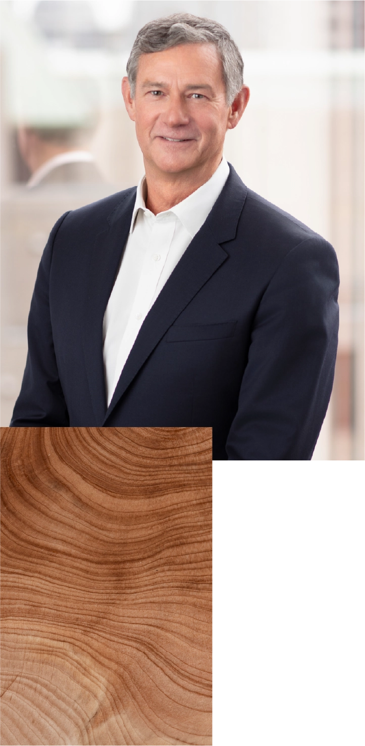 Une personne vêtue de manière professionnelle, portant un veston foncé et une chemise claire, se tient devant un arrière-plan intérieur légèrement flou. La personne est positionnée sur le côté gauche de l’image, faisant face vers l’avant. Du côté droit, un encadré rectangulaire présente un gros plan de grain de bois avec des cernes visibles et des tons bruns chaleureux.