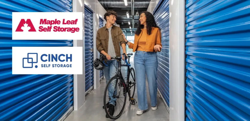 Two people walking a bicycle through a self-storage facility corridor lined with blue storage unit doors, with Maple Leaf Self Storage and Cinch Self Storage logos displayed on the left.