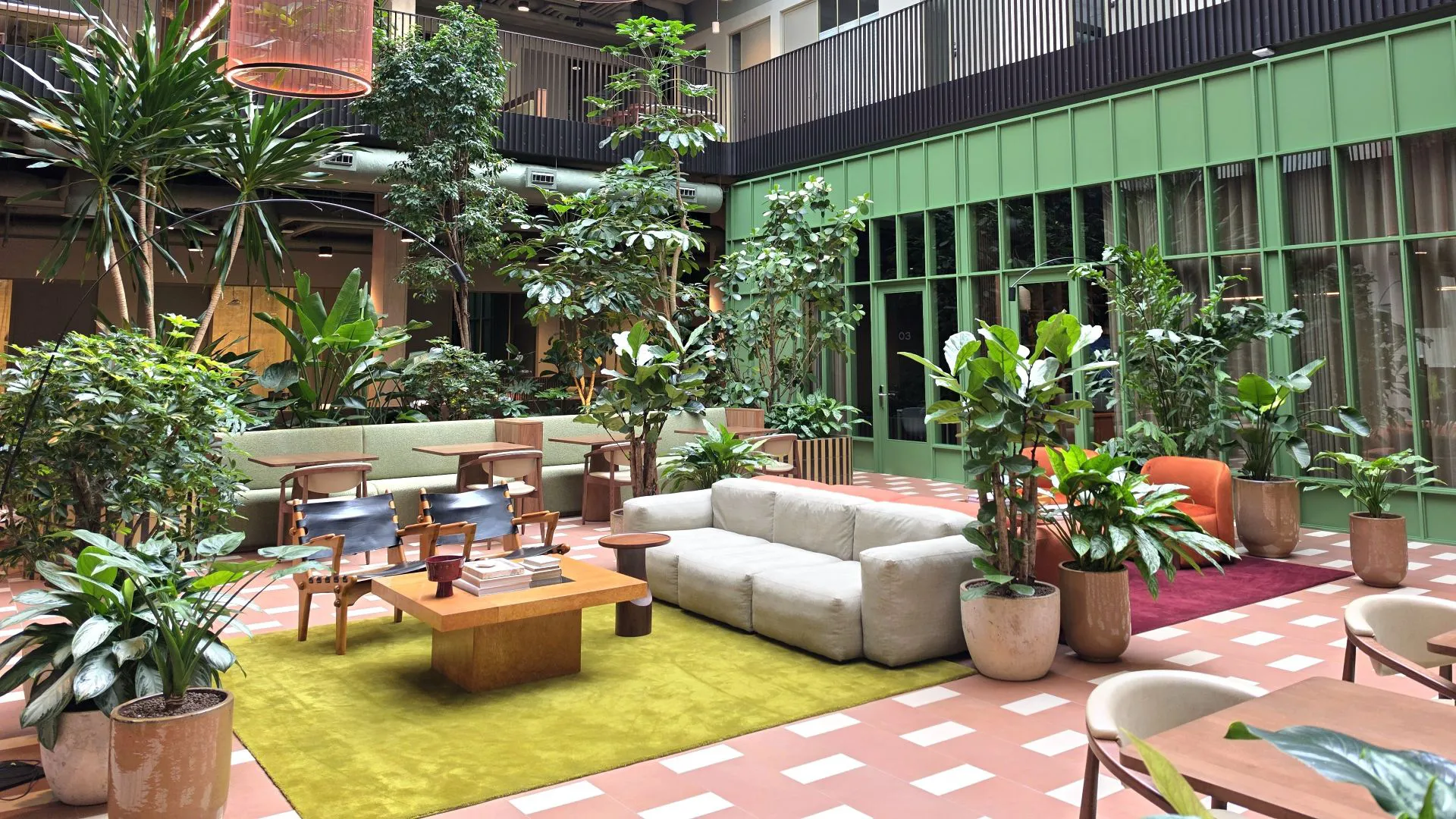 Modern indoor lounge filled with lush plants, featuring sofas, chairs, and tables arranged around a cozy seating area in a bright, garden-inspired atrium.