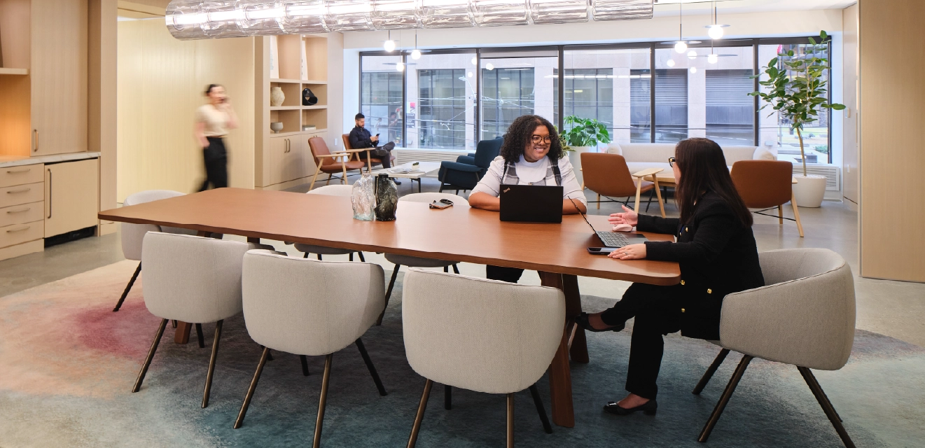 Modern office lounge and meeting space with a large communal table, seating areas, and people working on laptops in a bright, open interior.