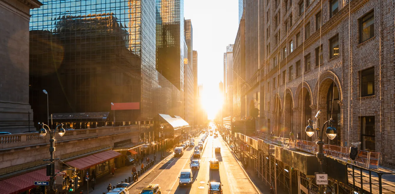 Sunlight streams down a busy downtown street lined with glass office towers and historic masonry buildings. Cars travel along the roadway as reflections of surrounding buildings glow in the windows during golden hour.