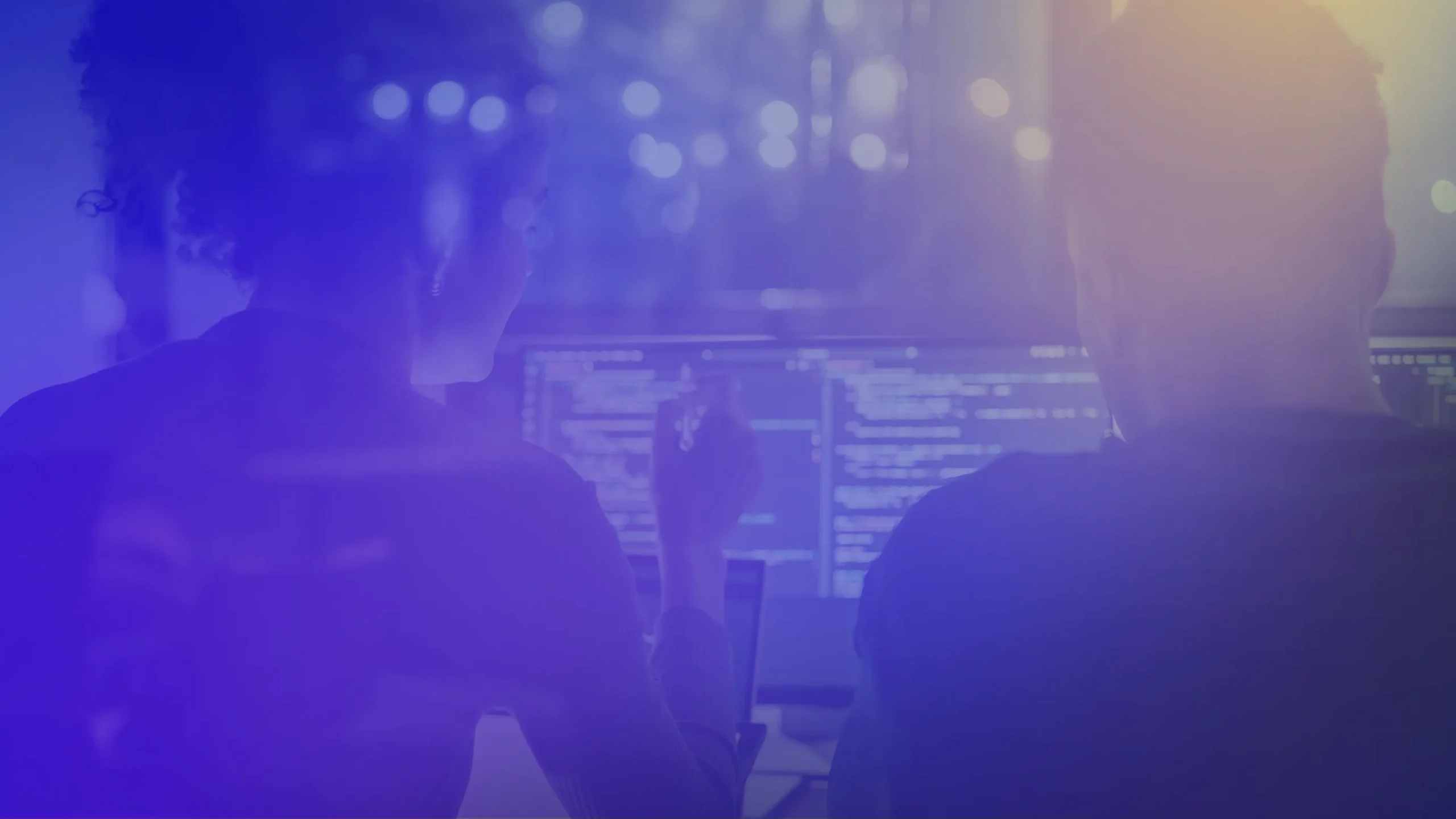 Two people seated at computer workstations, viewed from behind, looking at monitors displaying lines of code. The image features a blue and purple overlay with soft lighting effects and blurred background lights, creating a technology-focused and modern digital workspace scene.