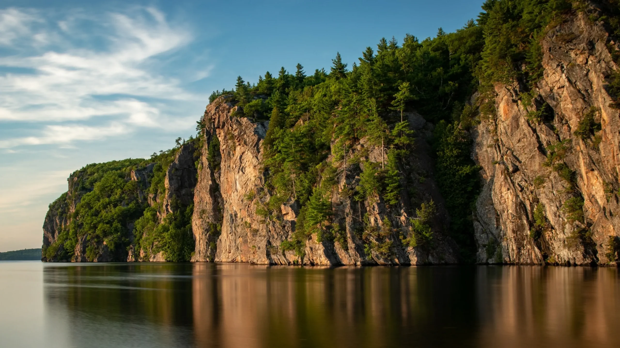 Steep rocky cliffs lined with evergreen trees rise above a calm body of water, reflecting the landscape below. Soft clouds drift across a blue sky, while warm sunlight highlights the rugged rock faces and dense forest.