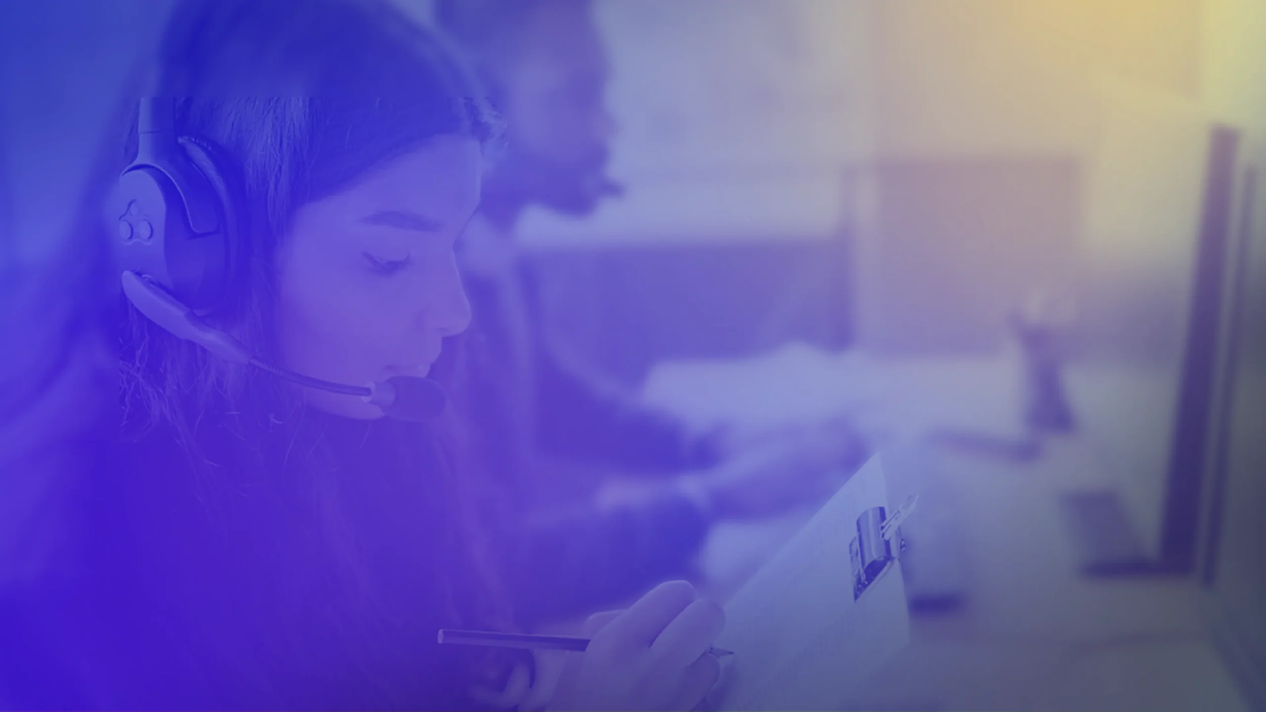 Close-up of a person wearing a headset and taking notes while working at a computer. A second headset-wearing colleague is visible in the background, with a blue-purple overlay across the image.
