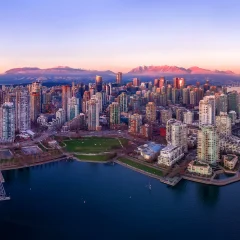 Aerial view of downtown Vancouver at sunrise, showing high‑rise buildings surrounded by water, marinas, and mountain ranges in the background.