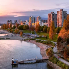 A scenic view of Vancouver’s waterfront at sunset, featuring a sandy beach, a tree-lined seawall, and high-rise residential buildings with mountains in the background.
