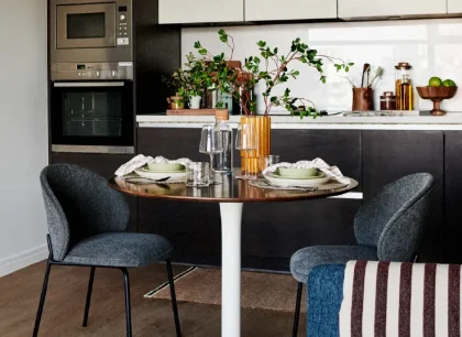 Contemporary apartment kitchen with integrated appliances, dark cabinetry, a round dining table for two and modern furnishings in an open-plan living space.