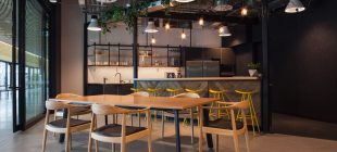 A modern kitchen and dining area with a wooden table and six chairs in the foreground. The kitchen features a long counter with four yellow bar stools, hanging pendant lights, and open shelves with glassware. There are plants hanging from the ceiling, adding a touch of greenery to the space.