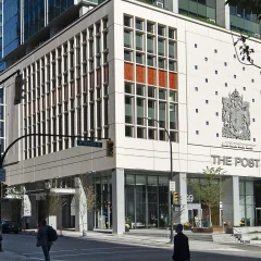 Street view of The Post building in downtown Vancouver, featuring a modern facade with vertical window panels, the building name ‘The Post’ and a large crest on the exterior wall. Pedestrians are crossing the intersection in front, and surrounding high-rise glass towers are visible in the background.