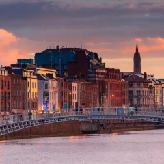 A scenic city view at sunset, featuring a pedestrian bridge over a river, colorful buildings along the waterfront, and historic spires in the background.