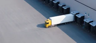 Aerial view of a yellow semi-truck with a white trailer parked at a loading dock of an industrial warehouse